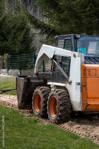 Obraz na płótnie A white and orange skid steer loader with a bucket attachment sits on dirt and grass. A green metal fence and tall evergreen trees are in the background. w salonie