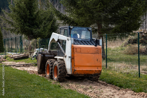 Obraz na płótnie An orange and white skid steer loader rests on a dirt path with its bucket down. The area features tire tracks, disturbed soil, and tall evergreen trees. w salonie