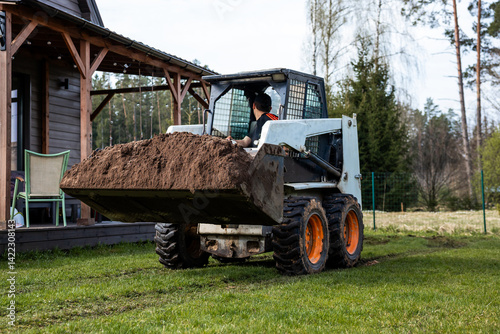 Obraz na płótnie A skid steer loader with orange wheels moves soil on grass near a wooden house with a covered porch, chairs, string lights, trees, and a fenced area. w salonie