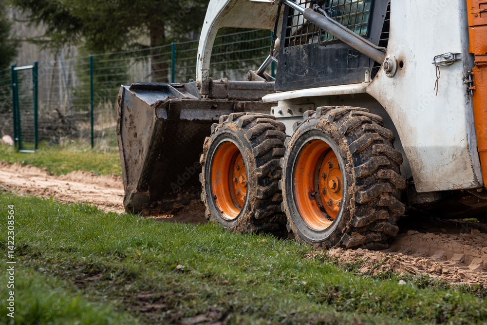 A compact skid steer loader with orange wheels and a front bucket rests on dirt and grass. Mud covered tires and a metal fence are visible nearby.