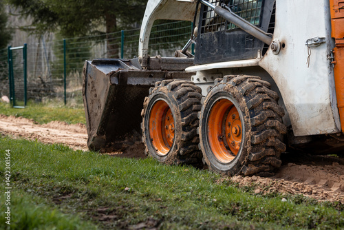 Obraz na płótnie A compact skid steer loader with orange wheels and a front bucket rests on dirt and grass. Mud covered tires and a metal fence are visible nearby. w salonie