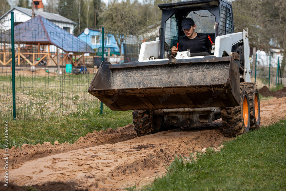A man uses a skid steer loader with a front bucket to level a dirt path. Green wire fence and residential houses with pitched roofs are visible nearby.