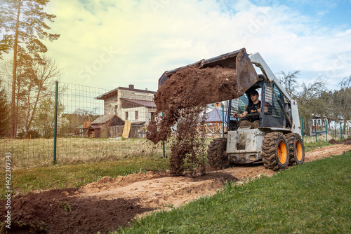 Obraz na płótnie A skid steer loader with orange wheels dumps soil on a pathway near a wooden house with a slanted roof, surrounded by fields and sparse trees. w salonie