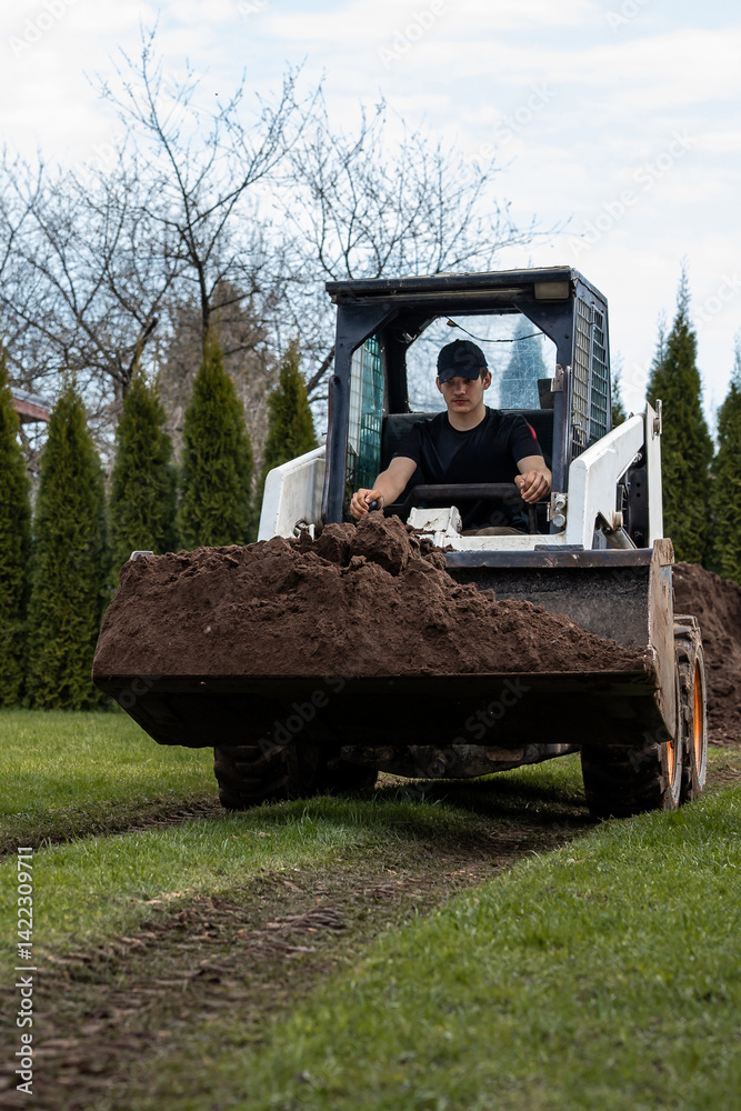 A man in a black cap and shirt operates a skid steer loader carrying soil across grass. Tracks are visible, with conifer and deciduous trees in the background.