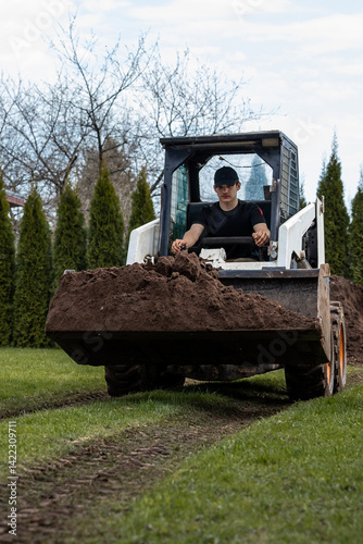 Obraz na płótnie A man in a black cap and shirt operates a skid steer loader carrying soil across grass. Tracks are visible, with conifer and deciduous trees in the background. w salonie