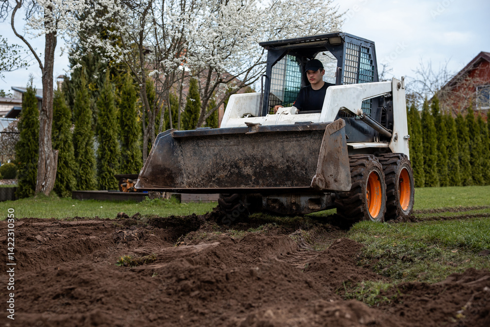A skid steer loader with orange wheels works on tilled soil. A person in a black cap operates it near flowering trees, hedges, and a red roofed house.