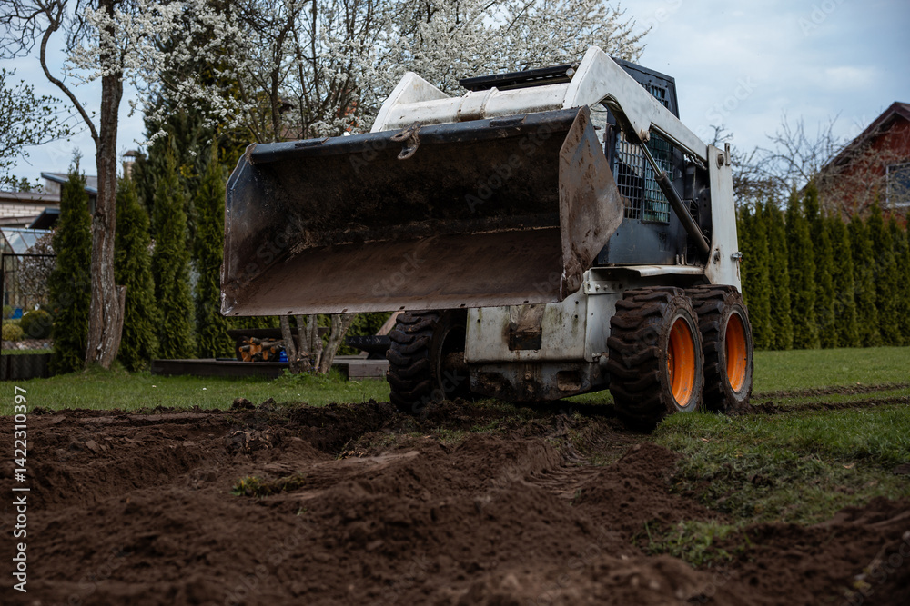 A skid steer loader with orange wheels rests on freshly dug soil near trees with white blossoms, trimmed hedges, and a house with a sloped roof.