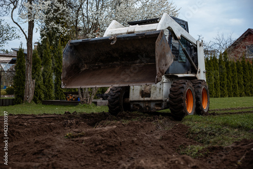 Obraz na płótnie A skid steer loader with orange wheels rests on freshly dug soil near trees with white blossoms, trimmed hedges, and a house with a sloped roof. w salonie