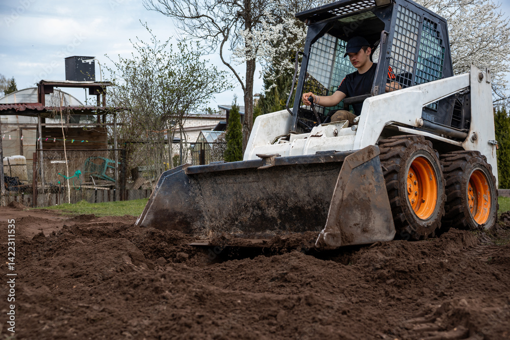 A man uses a skid steer loader with orange wheels to move soil. The area includes trees, some in bloom, a shed, fencing, and natural surroundings.