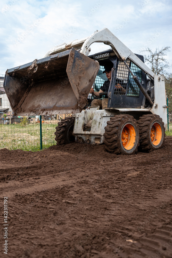 A skid steer loader with orange wheels raises a bucket filled with soil. The scene shows freshly tilled ground, a wire fence, and distant structures.