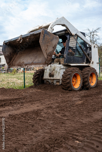 Obraz na płótnie A skid steer loader with orange wheels raises a bucket filled with soil. The scene shows freshly tilled ground, a wire fence, and distant structures. w salonie