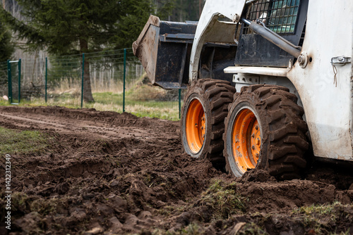 A skid steer loader with orange wheels sits on a muddy dirt path, its bucket raised. A green metal fence and sparse vegetation are visible nearby.