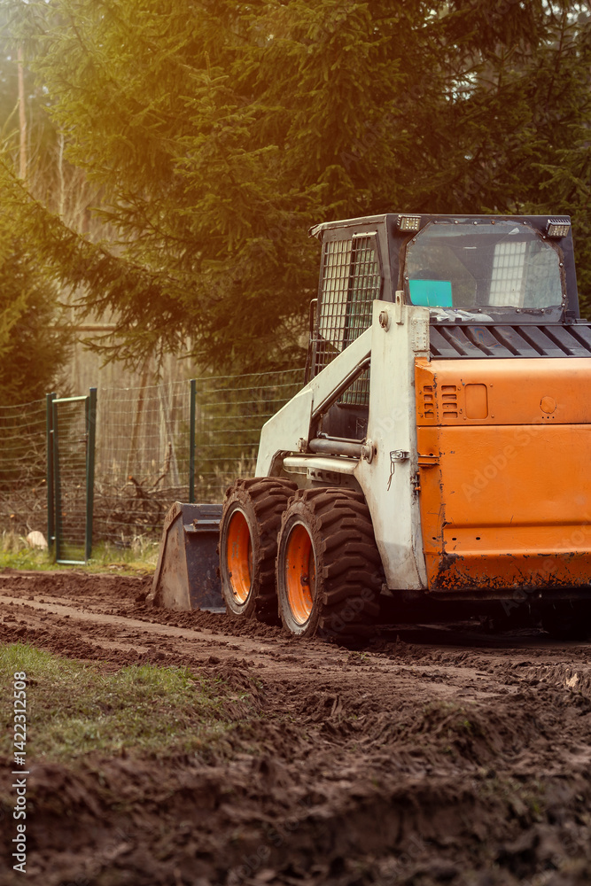 An orange and white skid steer loader rests on a muddy path with its bucket down, surrounded by a wire fence and tall evergreen trees in sunlight.