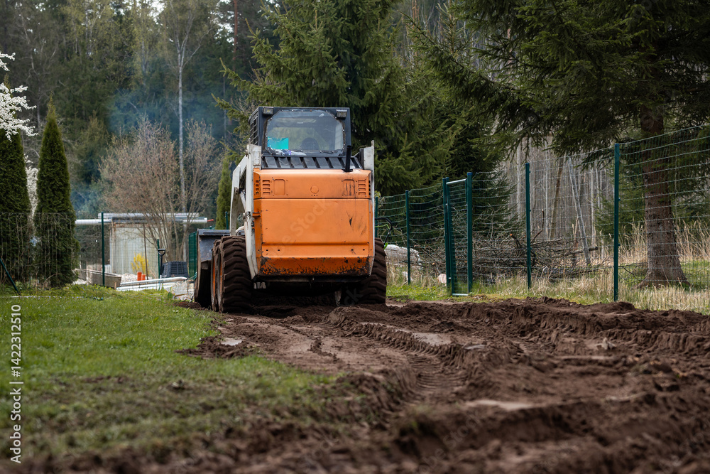 An orange and white skid steer loader works on a dirt path, leaving tire tracks. A green metal fence, tall evergreen trees, and a structure with a transparent roof are visible.
