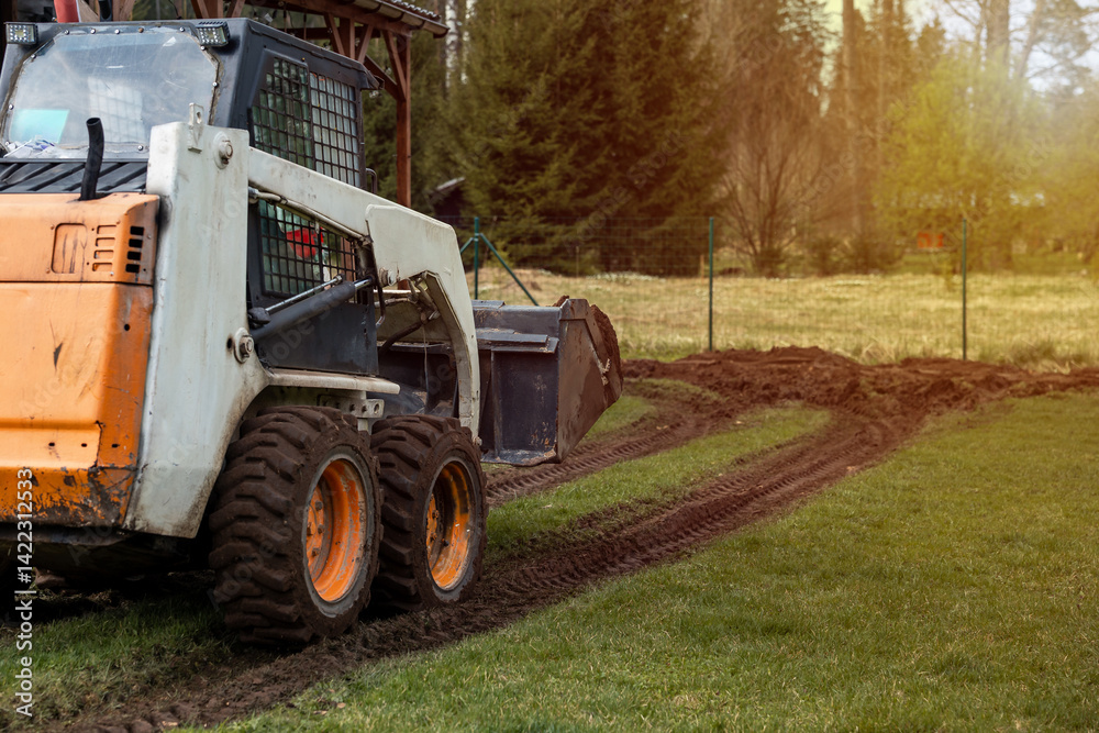 A compact orange and white skid steer loader moves across a grassy field, leaving tire tracks. A wire fence and tall trees are visible in the distance.