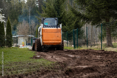 Obraz na płótnie An orange and white skid steer loader works on a dirt path, leaving tire tracks. A green metal fence, tall evergreen trees, and a structure with a transparent roof are visible. w salonie