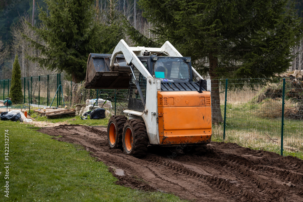 A compact orange and white skid steer loader operates on a dirt path, leaving tire tracks. Surrounded by a wire fence, evergreen trees, logs, and bags.