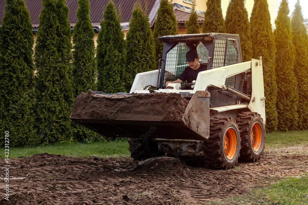 A skid steer loader with a raised bucket moves soil near tall evergreen trees. A building with a sloped roof is partially visible in the setting.