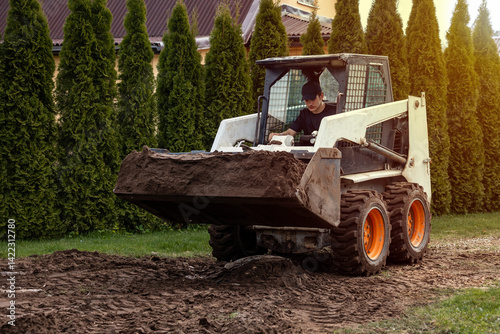 Obraz na płótnie A skid steer loader with a raised bucket moves soil near tall evergreen trees. A building with a sloped roof is partially visible in the setting. w salonie