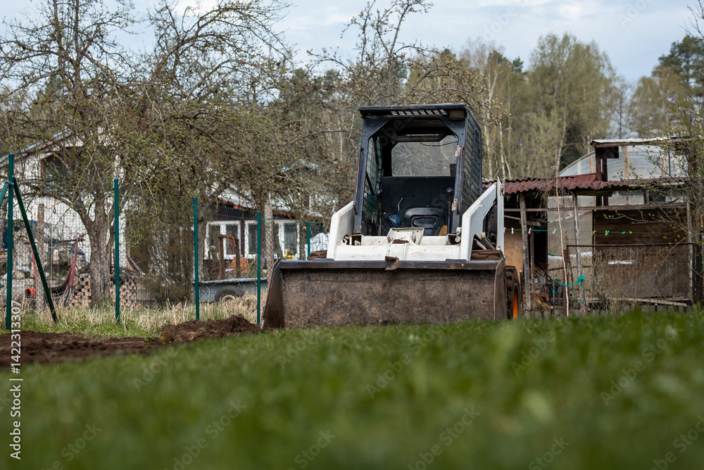 A white skid steer loader with its bucket lowered sits on grass, surrounded by sparse trees, a fenced area, a shed, and a house in a rural setting.