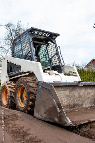 Obraz na płótnie A skid steer loader with a front bucket sits on muddy ground. Leafless trees and a red roofed building partially hidden by shrubs are visible. w salonie