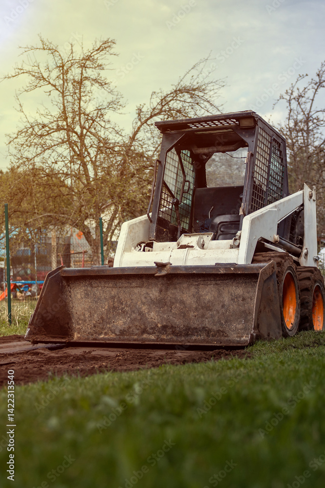 A white skid steer loader with orange wheels sits on a dirt patch surrounded by grass. Leafless trees and a faint fence are visible in the background.
