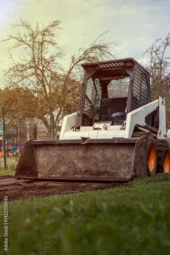 Obraz na płótnie A white skid steer loader with orange wheels sits on a dirt patch surrounded by grass. Leafless trees and a faint fence are visible in the background. w salonie