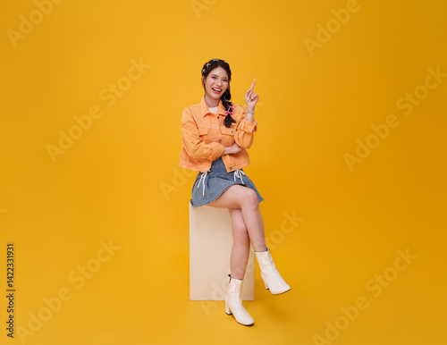 Happy cute Asian teen girl sitting on chair and relax hand pointing finger up isolated on yellow  background. People lifestyle concept.