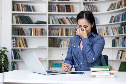 An asian woman rubs her tired eyes while studying at a library. She sits at a desk with a laptop, glasses, and books.
