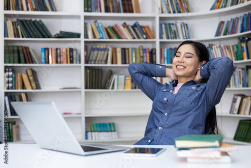 A relaxed woman smiles with her eyes closed, enjoying a break in the library. Her arms rest behind her head as she sits at her desk. Bookshelves fill the background.