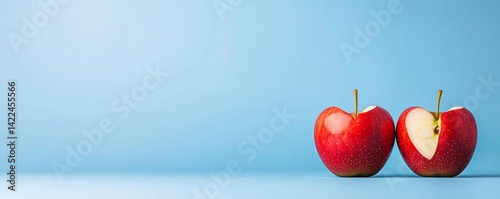 Two ripe red apples are sitting side by side, one with a bite taken out of it, against a solid pastel blue background for concepts.