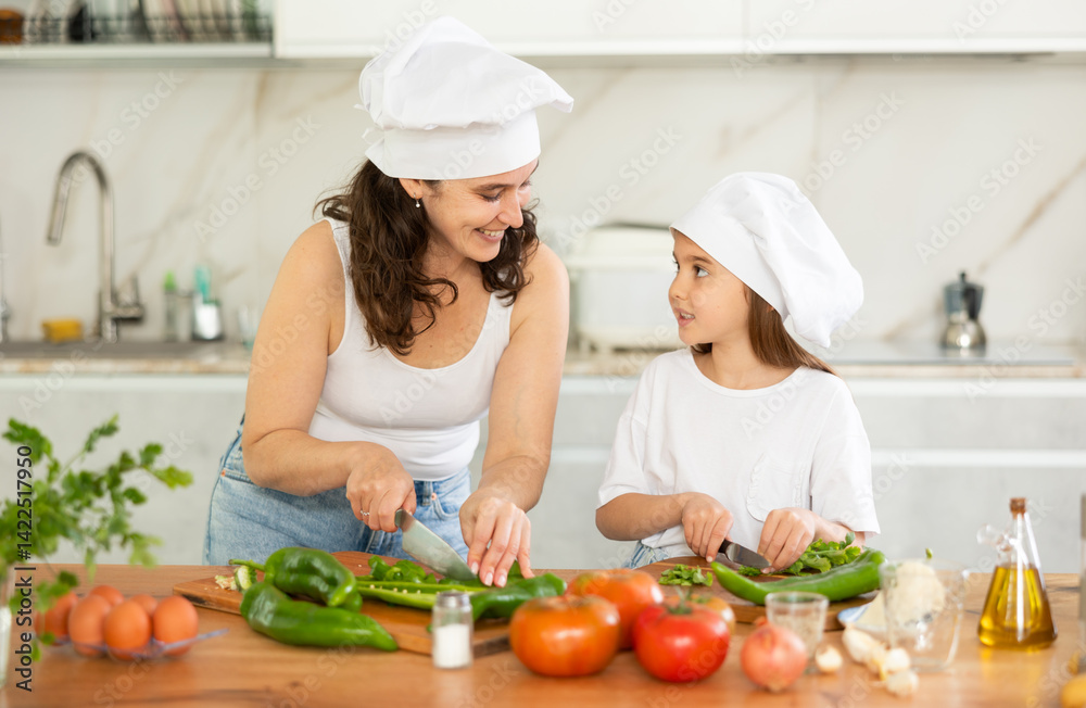 Mom and daughter in hats dice up raw vegetables for salad on cutting boards. Girl listens attentively to her mother, carefully works with knife, tries to follow recommendations Mom and daughter in hats dice up raw vegetables for salad on cutting boards. Girl listens attentively to her mother, carefully works with knife, tries to follow recommendations