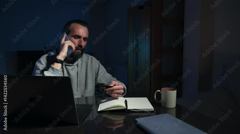 Man Working From Home on Laptop at Night, Talking on Phone and Taking Notes Man Working From Home on Laptop at Night, Talking on Phone and Taking Notes
