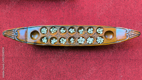 Top-down view of a traditional Congklak board game with seeds placed in carved wooden pits, set against a red textured background.