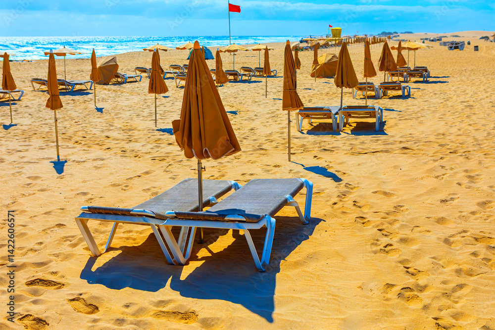 Beach scene with rows of lounge chairs and umbrellas facing ocean, all neatly arranged on sandy shore. Red flag in background signals hazardous swimming conditions, and beach is completely unoccupied