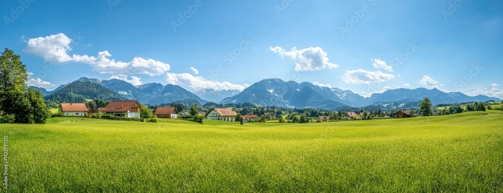 Fototapeta premium Idyllic village nestled in a green valley, majestic mountains in the background under a bright blue sky.