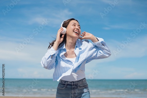 A joyful young Asian woman listening to music with headphones at the beach, smiling brightly under the clear blue sky.enjoying and relaxing summer atmosphere concept.