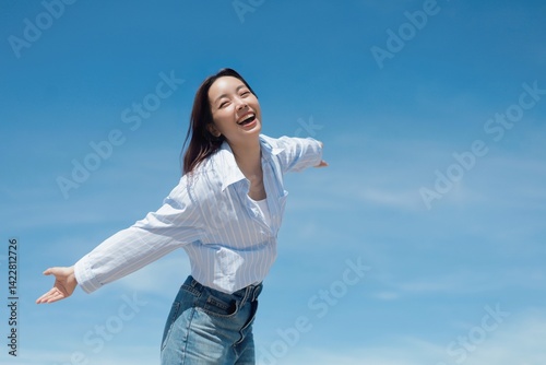 A cheerful Asian woman enjoying a sunny day at the beach. She is wearing a casual shirt and jeans standing in front of the blue sky background