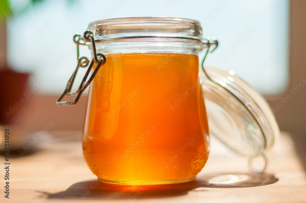 A jar filled with golden honey sits on a light wooden table. Sunlight pours in, highlighting its warm tones and inviting sweetness, perfect for culinary use.