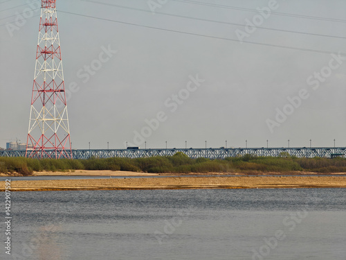 A tall lattice power line tower stands near a calm river, with a bridge stretching across the water. the scene is framed by thin vegetation along the riverbank, set against a clear sky. the intricate