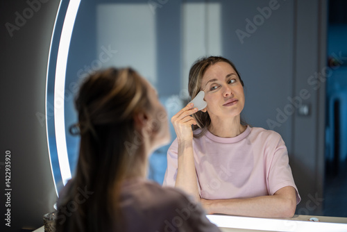 Calm mindful middle aged woman enjoying self-care morning routine, gently massaging face with gua sha stone. Skincare ritual of focused mature female with natural beauty aging. Harmony with self love. photo