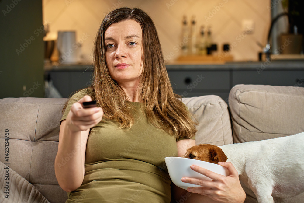 Woman relaxing on sofa with remote control while dog steals food. Pet dog eat food from bowl while owner focuses on TV. Home life and pet behavior concept