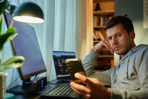 Obraz na płótnie Tired freelancer procrastinating at home office, scrolling phone instead of working. Unproductive man using smartphone at home desk. Concept of procrastination and distraction in remote work w salonie