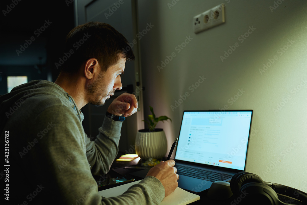Concentrated freelancer working late at home office. Focused man in hoodie writes notes while working on laptop. Concept of night work