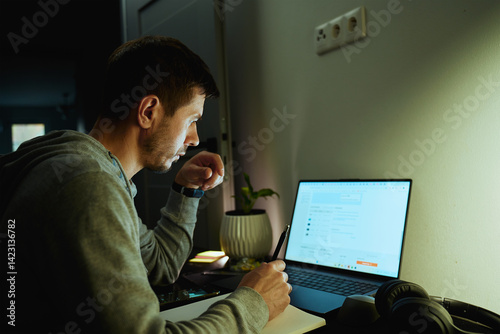 Obraz na płótnie Concentrated freelancer working late at home office. Focused man in hoodie writes notes while working on laptop. Concept of night work w salonie