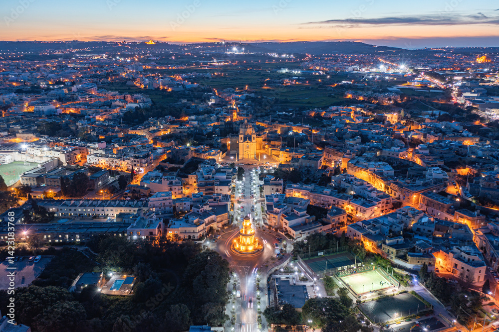 Fototapeta premium Panoramic view of Lija village in Malta. The Belveder Tower and Parish Church are lit. Mdina can be see in the distance during just after sunset.