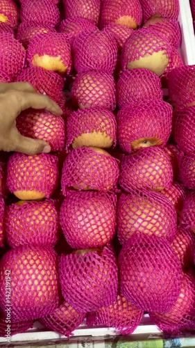A man's hand selects a red apple with a white wrapper on a fruit vendor's table.