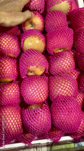A man's hand selects a red apple with a white wrapper on a fruit vendor's table.