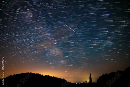 Star trails of a Milky Way with meteor, shooting stars and countryside silhouettes.