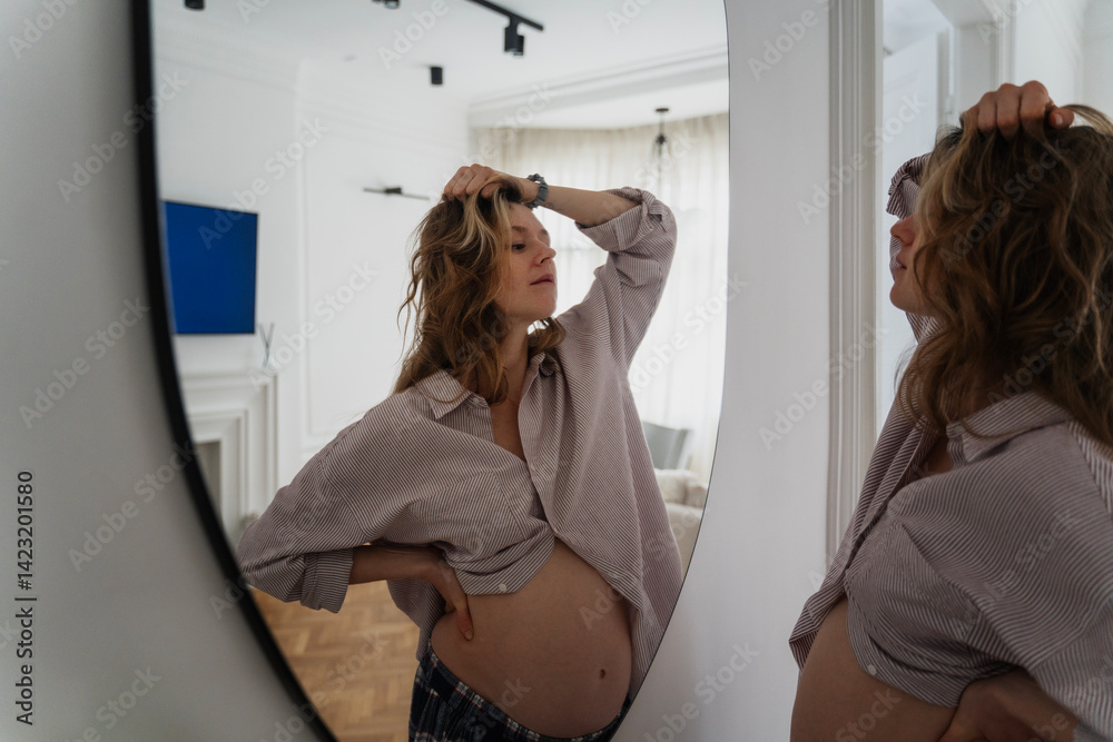 Young attractive pregnant woman looking at herself in the mirror while spending time at home